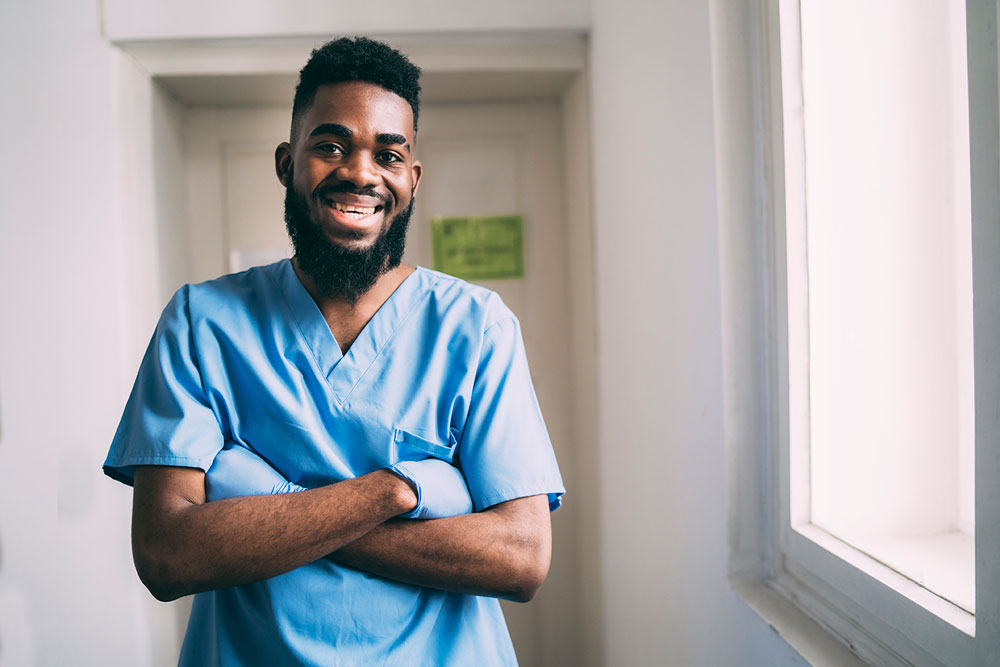 Smiling doctor standing with his arms crossed wearing protective gloves and hospital scrubs in a hospital corridor